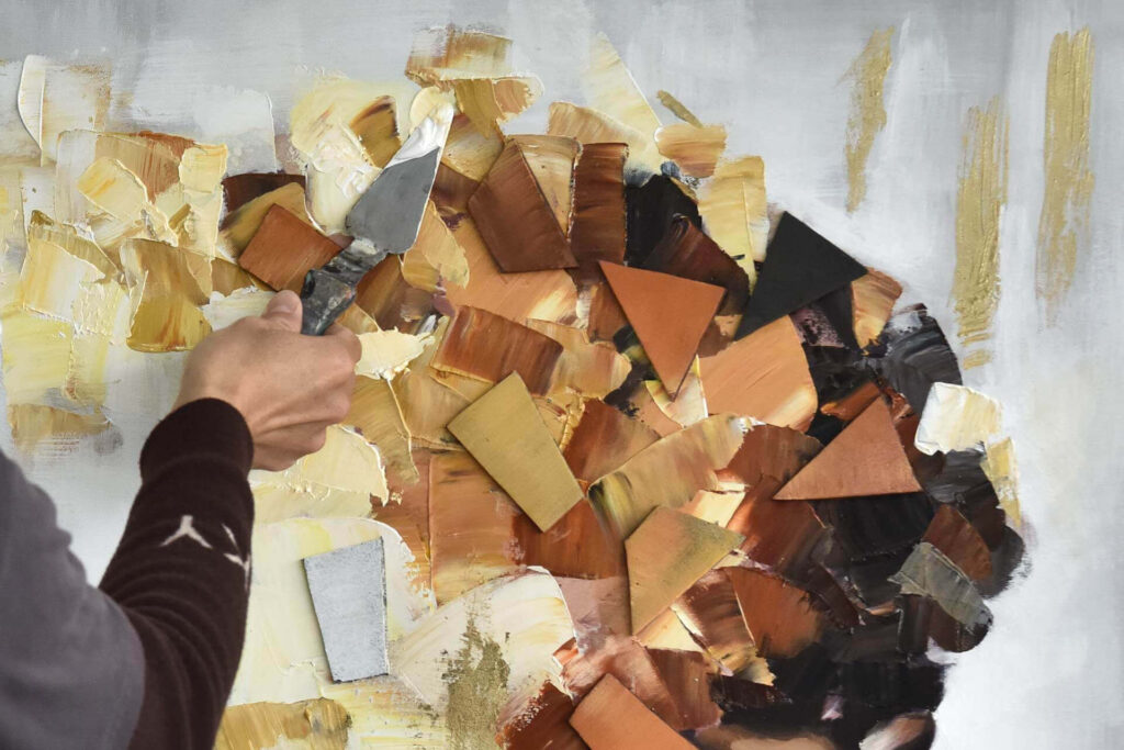 A close-up shot showing an artist using a palette knife to apply thick, textured brown, gold, and cream paint layers onto a canvas.